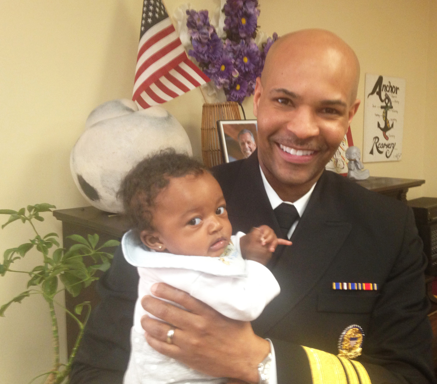 U.S. Surgeon General Dr. Jerome Adams holding a young baby girl during an interview at the Anchor Recovery Community Center on Jan. 26. Above his shoulder is the photograph of a smiling Jim Gillen, one of the leaders of the recovery community in Rhode Island.